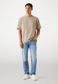 Beige katoenen t-shirt met een ronde hals en een klein voorzakje, gecombineerd met lichtblauwe denim jeans en witte sneakers.