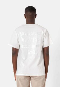 Camiseta de algodón blanca con un gráfico texturizado en la parte posterior que dice "Los miedos que no enfrentamos se convierten en nuestros límites." Mangas cortas.