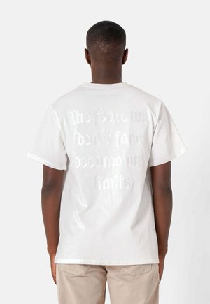 Weißes Baumwoll-T-Shirt mit einem strukturierten Grafikdesign auf der Rückseite, das den Text "Die Ängste, denen wir uns nicht stellen, werden zu unseren Grenzen." trägt. Kurze Ärmel.