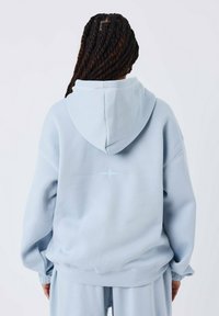 Persona con capelli intrecciati che indossa un grande felpa azzurro chiaro con un piccolo design a stella sulla schiena, abbinata a pantaloni coordinati.