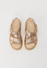 Sandales en bronze avec des sangles croisées et une boucle, présentant un design cousu et une semelle texturée pour une meilleure adhérence et un confort optimal.