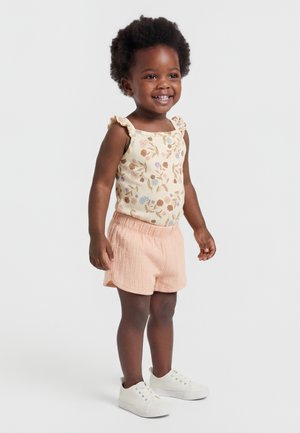 Χαμογελαστή μικρή κοπέλα με σγουρά μαλλιά, φοράει φλοράλ αμάνικο τοπ, ροδακινί σορτσάκι και λευκά αθλητικά παπούτσια, στέκεται σε λευκό φόντο.