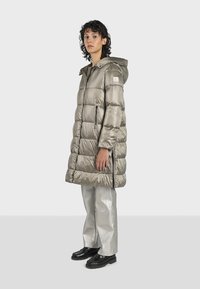 Cappotto lungo imbottito in tessuto grigio metallico con cappuccio, zip frontale e tasche laterali. Abbinato a pantaloni argentati lucidi e scarpe nere.