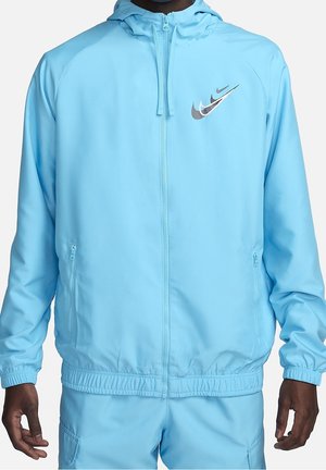 Homme portant un sweat à capuche zippé Nike bleu clair et un short assorti, avec des logos Nike swoosh superposés sur la poitrine.