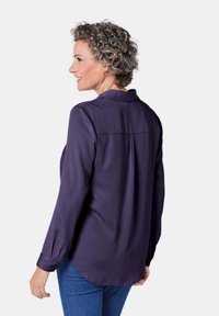 Purple long-sleeve shirt with a collar, buttoned cuffs, and back pleat detail, paired with blue jeans. Smooth texture, relaxed fit.