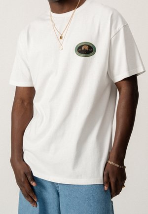 Mężczyzna w białym krótkim rękawem t-shircie z zielono-złotym logo drzewa na piersi, niebieskich dżinsach, złotych naszyjnikach i bransoletce.