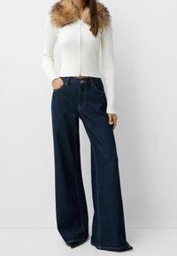 Vit ribbad zip-up kofta med en brun pälsöverkant, i kombination med mörkblå vida jeans. Noterbar sömnad och framfickor synliga.