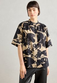 Kortermet skjorte med knapper i beige bomull med et svart blomstermønster. Har en stående krage og rynker langs skulderpartiet.