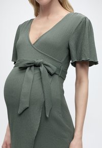 Έγκυος γυναίκα που φοράει υφασμάτινο φόρεμα τύπου wrap σε χρώμα φιδιού πρασινωπό, με κοντά μανίκια που ανεμίζουν και δεμένο φιόγκο στη μέση.