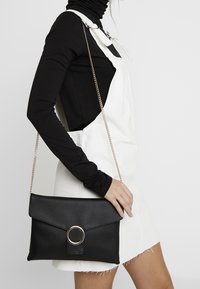 Svart konstläder crossbody-väska med en vikbar klaff, cirkulär roséguldaccent och kedjestrat, hålls av en person iklädd en svart topp.