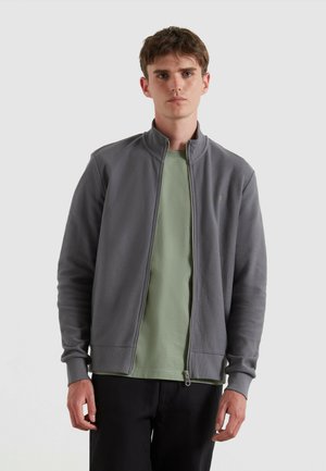 Veste à zip grise avec une surface texturée et un col haut, portée sur un t-shirt vert clair, associée à un pantalon sombre.