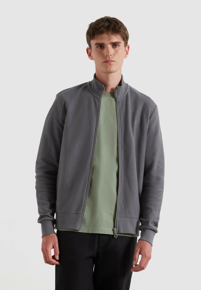Grijze zip-up jack met een gestructureerd oppervlak en hoge kraag, gedragen over een lichtgroene t-shirt, gecombineerd met donkere broeken.