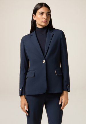 Blazer blu navy realizzato in tessuto liscio, con un singolo bottone dorato, due tasche frontali e vestibilità su misura. Leggermente strutturato con un rever classico.