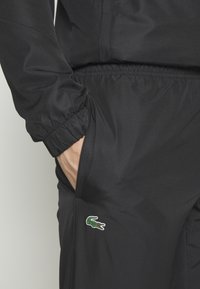 Čierna atletická bunda s elastickými manžetami, zipsovým vreckom a zeleno-červeným logom krokodíla Lacoste na ľavej strane.