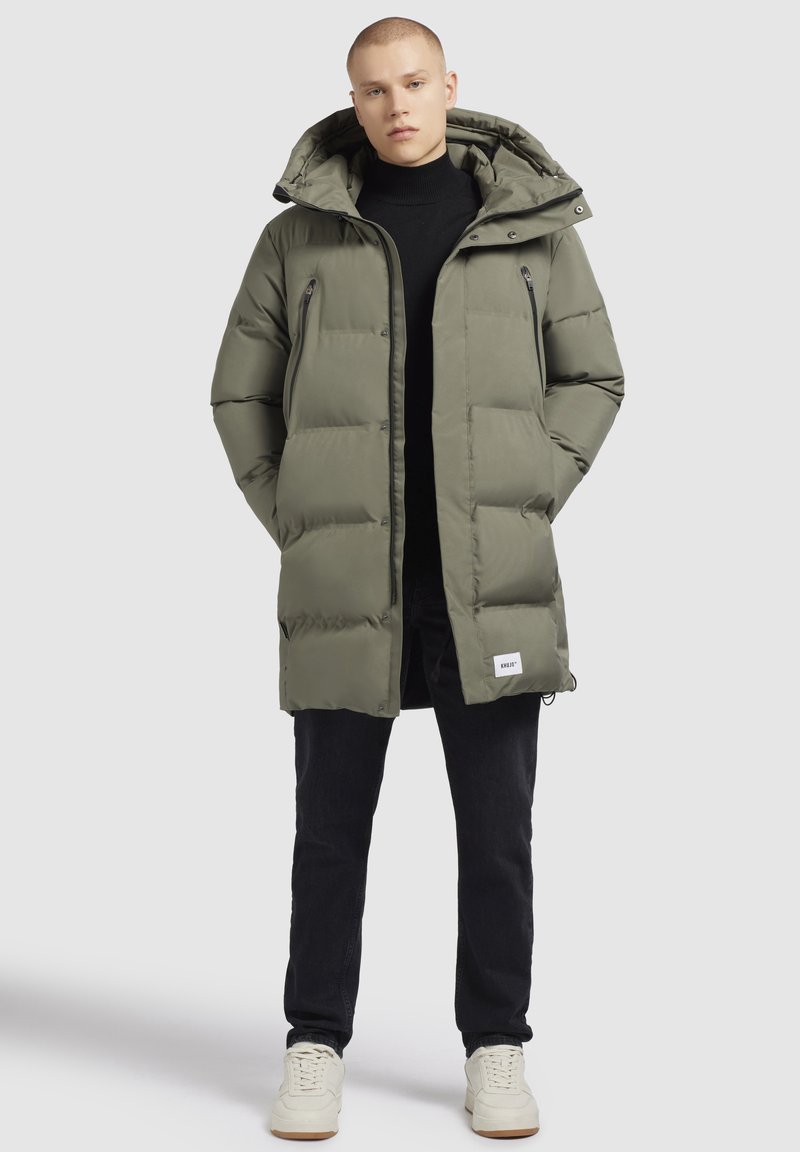 Parka căptușit de culoare verde măslin, cu glugă, buzunare cu fermoar și manșete elastice, purtat cu un pulover negru și blugi închiși la culoare.