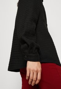 Zwarte textuurtrui met een relaxte pasvorm, voorzien van een knoopmanchet en een ronde zoom. Draagt over rode broek, wat de kleurcontrasten accentueert.