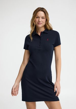 Femme aux cheveux blonds ondulés portant une robe polo bleu marine à manches courtes avec des boutons et un petit logo rouge brodé sur la poitrine.