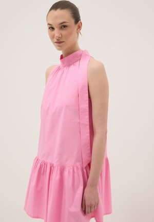 Frau mit zurückgebundenem Haar, die ein ärmelloses rosa Kleid mit hohem Ausschnitt und einer gebundenen Schleife auf der linken Schulter vor einfachem Hintergrund trägt.