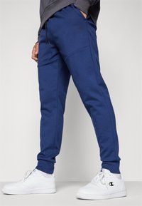 Blaue Sweatpants aus einem weichen Baumwoll-Mix, mit einem Kordelzugbund, seitlichen Taschen und gerippten Bündchen. Tragen mit weißen Sneakern.