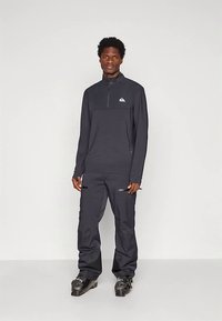 Man in een zwarte ritssportjack, zwarte broeken en skibotten, staande tegen een effen lichte achtergrond.