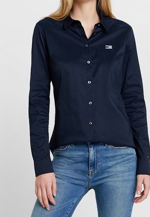 Mujer con una camisa azul marino abotonada de manga larga y vaqueros azules, de pie con una mano apoyada en su muslo.