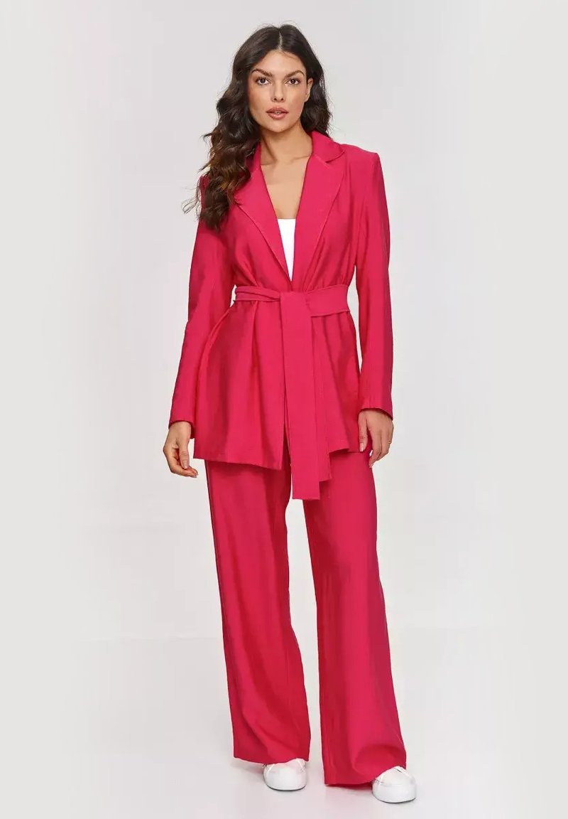 Jasnoróżowy, dopasowany garnitur z długim, otwartym żakietem i szerokimi spodniami. Posiada paskowane wcięcie w talii oraz gładką teksturę.