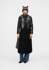 Νεαρό άτομο που φοράει μαύρο δερμάτινο μπουφάν πάνω από μαύρο πλισέ φόρεμα με μπλε τζιν, κρατώντας τσάντα με καρφιά και ριγέ πλεκτό καπέλο με αυτιά.