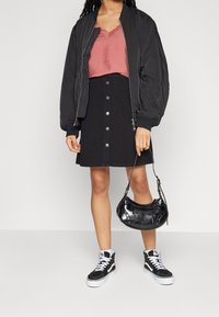 Woman wearing a black bomber jacket over a pink lace-trim camisole, black button-front skirt, black and white high-top sneakers, holding a black handbag.