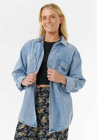 Jasnoniebieska koszula z denimu z klasycznym kołnierzykiem, przednimi kieszeniami i zapięciem na guziki, noszona rozpięta na czarnym topie crop i spódnicy w kwiatowy wzór.