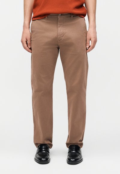 Homme portant un pantalon marron droit, des chaussures habillées noires et un pull texturé orange, debout devant un fond blanc.