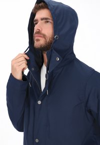 Marineblaue wasserdichte Jacke mit Kapuze, Reißverschluss und Druckknöpfen, strukturiertem Stoff und silbernen Beschlägen.