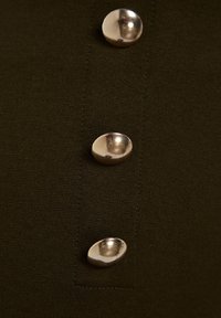 Trois boutons métalliques brillants sur un tissu sombre texturé, disposés verticalement, avec des coutures visibles autour des boutons.