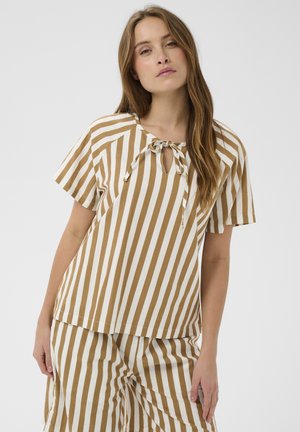 Kvinde med langt brunt hår iført tan og hvidt vertikalt stribet kortærmet top og matchende bukser mod en ensfarvet baggrund.