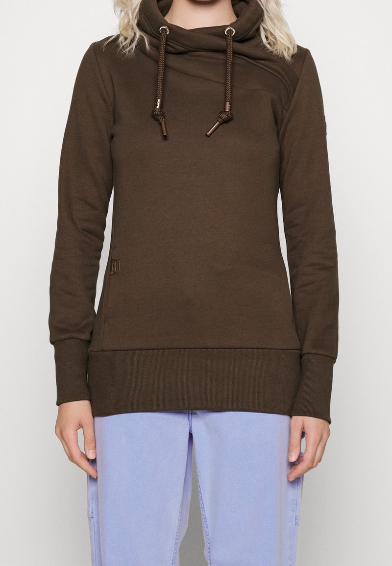 Femme portant un sweat à capuche marron à manches longues avec des cordons et un pantalon bleu clair, debout devant un fond uni.