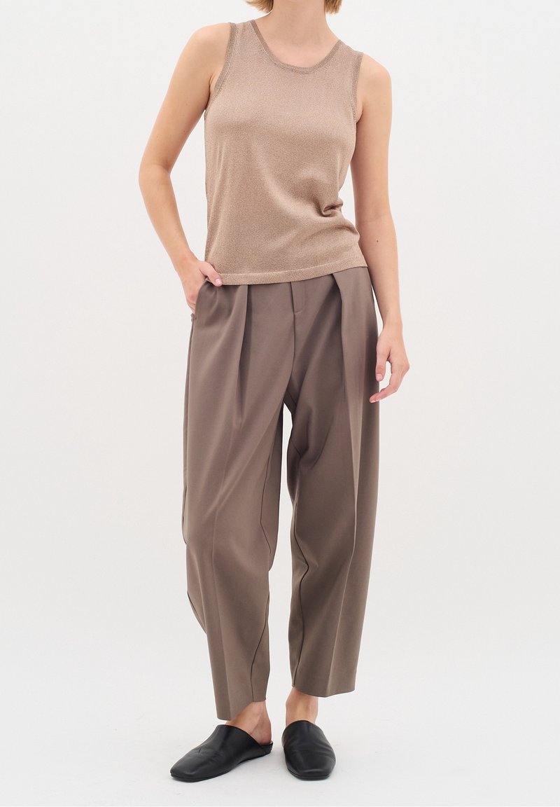 Femme portant un haut beige sans manches, un pantalon plissé marron ample et des chaussures noires à enfiler, debout avec une main dans la poche.