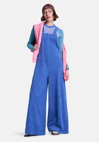 Μπλε βελούδινη φόρμα με φαρδιά πόδια, ρυθμιζόμενους ιμάντες και υφή επιφάνειας. Ένα ροζ φούτερ είναι περασμένο πάνω σε έναν ώμο.