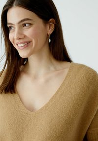 Hellbrauner, flauschiger Pullover mit einem tiefen V-Ausschnitt und kurzen Ärmeln. Model trägt Perlenohrringe, hat helle Haut und lange Haare.