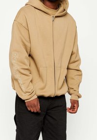 Felpa con zip beige realizzata in tessuto morbido, dotata di tasca a marsupio, polsini a costine e design stampato lungo la manica sinistra.