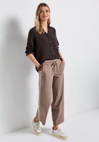 Brun skjorta med knappar och uppvikta ärmar, kombinerad med beige vida byxor med sidstripes. Modellen har på sig vita sneakers.