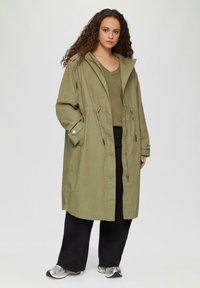 Olijfgroene lange jas met een capuchon, verstelbare taille en zijzakken. Gepaard met een groene top en zwarte broek. Zilveren sneakers zichtbaar.