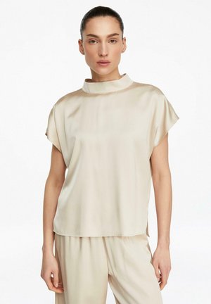 Frau trägt eine lockere, kurzärmelige, hochgeschlossene beige Satinbluse und passende Hose vor weißem Hintergrund.