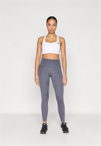 Brassière de sport blanche avec logo noir, leggings taille haute gris et baskets noires. Tissu lisse, coupe ajustée, sans motifs ni accents visibles.