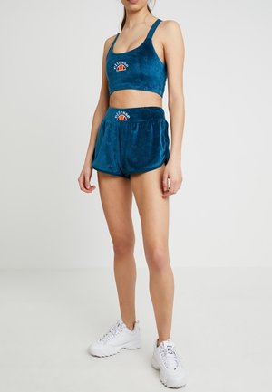 Modèle féminin portant un crop top Ellesse en velours bleu et un short assorti avec des baskets blanches, debout devant un fond clair uni.