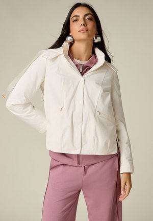 Kvinde iført en hvid kort jakke over en mauve lynlåstop og matchende mauve bukser, stående med den ene hånd på hoften mod en ensfarvet baggrund.