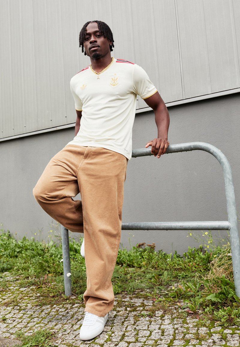 Cremefarbiges Hemd mit roten Streifen und goldenen Akzenten, kombiniert mit braunen Cordhosen und weißen Sneakers. Ein Mann lehnt an einem Geländer.