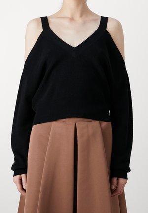 Person, die einen schwarzen Cold-Shoulder-Pullover mit langen Ärmeln trägt, kombiniert mit einem hoch taillierten, plissierten braunen Rock.