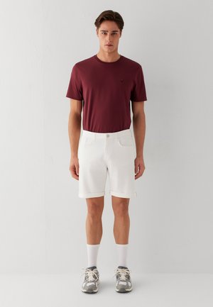 Junger Mann steht, trägt ein burgunderrotes T-Shirt, weiße Shorts, weiße Socken und graue Sneakers vor einem einfarbigen hellen Hintergrund.