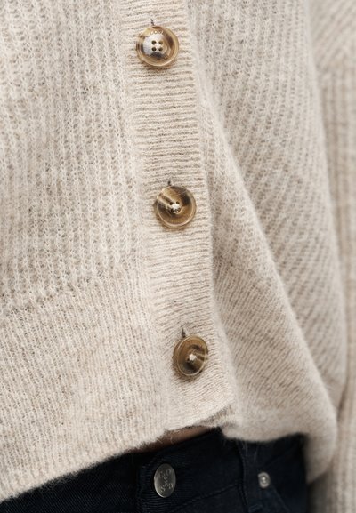 Μπεζ πουλόβερ πλεκτό ζακέτα με μεγάλα καφέ κουμπιά πάνω από σκούρο μπλε τζιν, που δείχνει υφή και λεπτομέρειες κουμπιών σε κοντινό πλάνο.