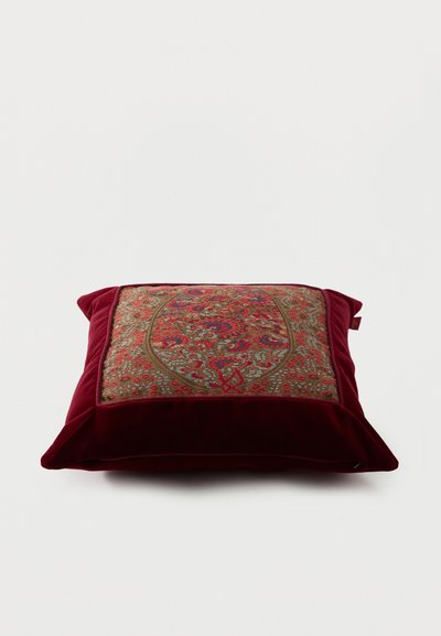 Coussin décoratif carré avec un motif floral rouge et or complexe sur le devant et une bordure en velours rouge profond sur un fond blanc.