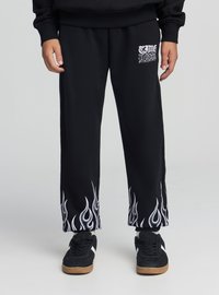 Czarne spodnie dresowe z białymi grafikami płomieni na kostkach. Materiał ma miękką teksturę. Z przodu na górnej części uda znajduje się małe logo.